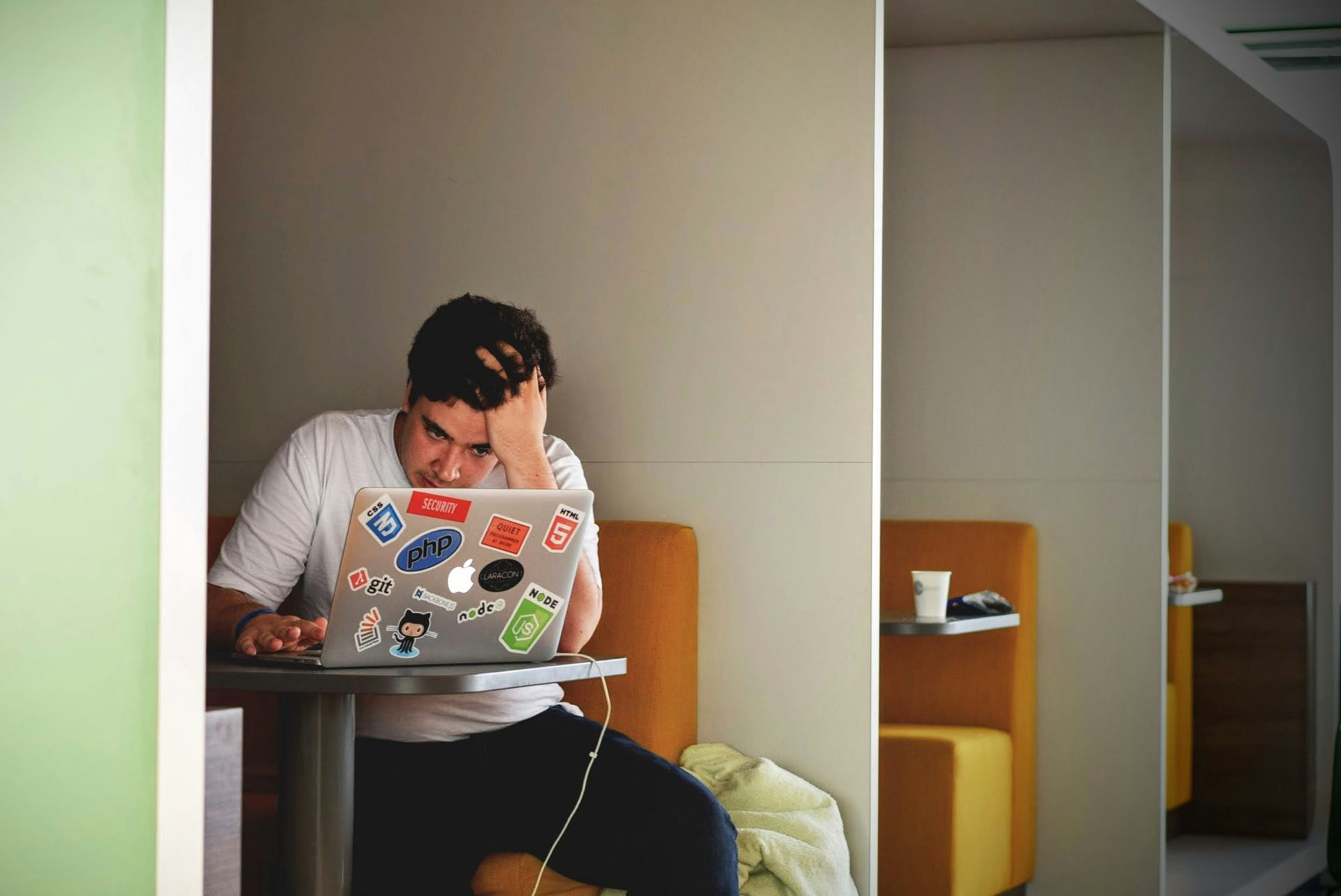 a man sat at a table at a diner holding his head with one hand.  He is looking anxiously at a laptop computer.