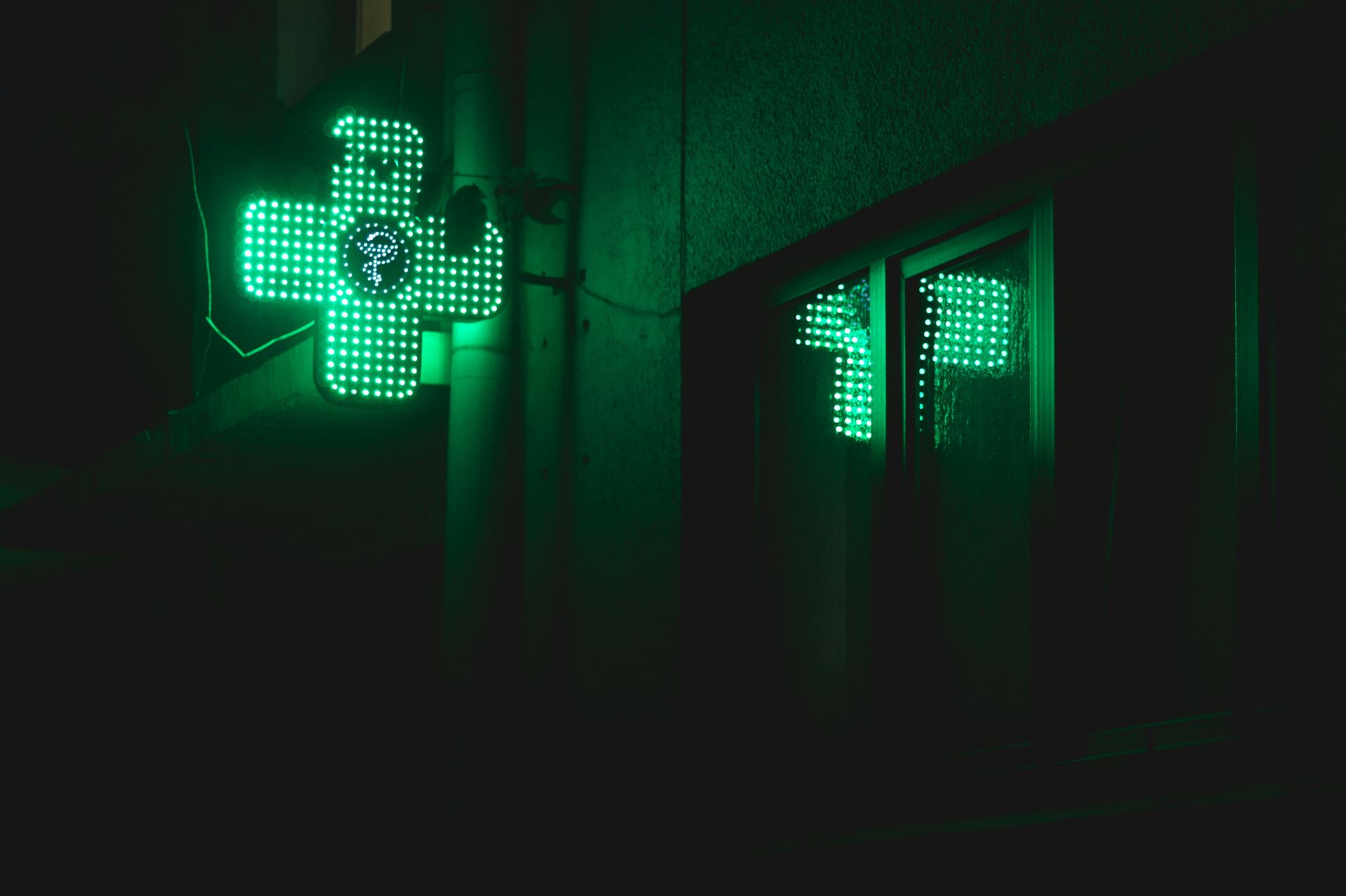 an green neon pharmacy sign shaped like a cross and reflected in a shop window