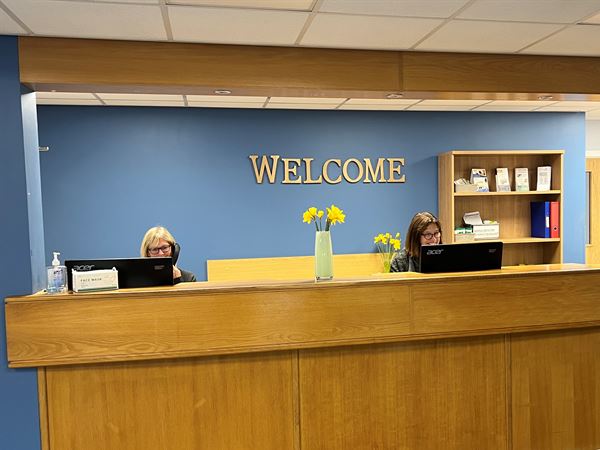 Verwood Surgery Reception counter with two receptionists, one is on the phone with a patient, the other is working on a computer.  There is vase of flowers on the counter and a sign saying "welcome" in yellow letters on a blue wall behind. 