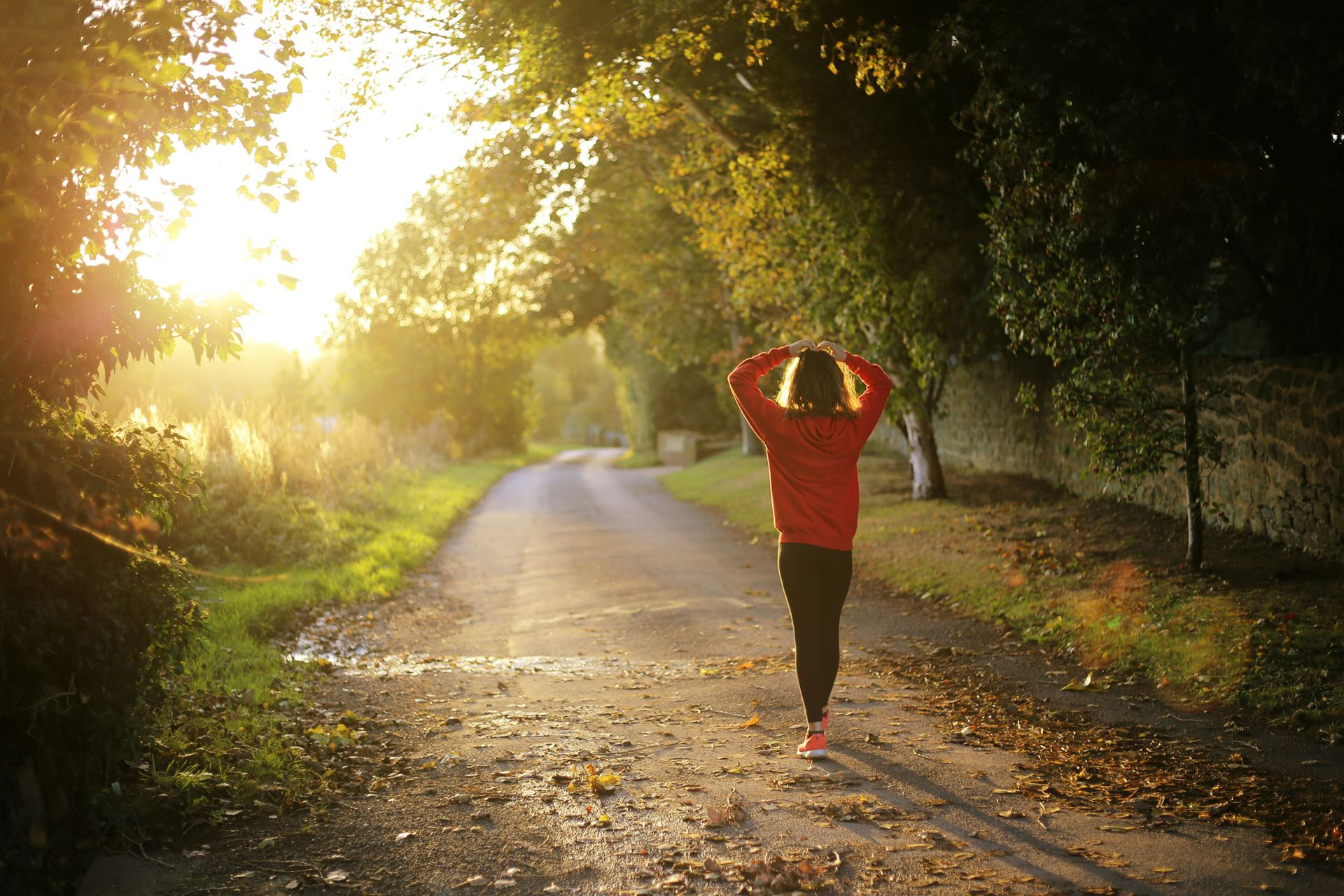 a woman walking in a park, the sun rising in front of her.  She has her hands in the air after jogging
