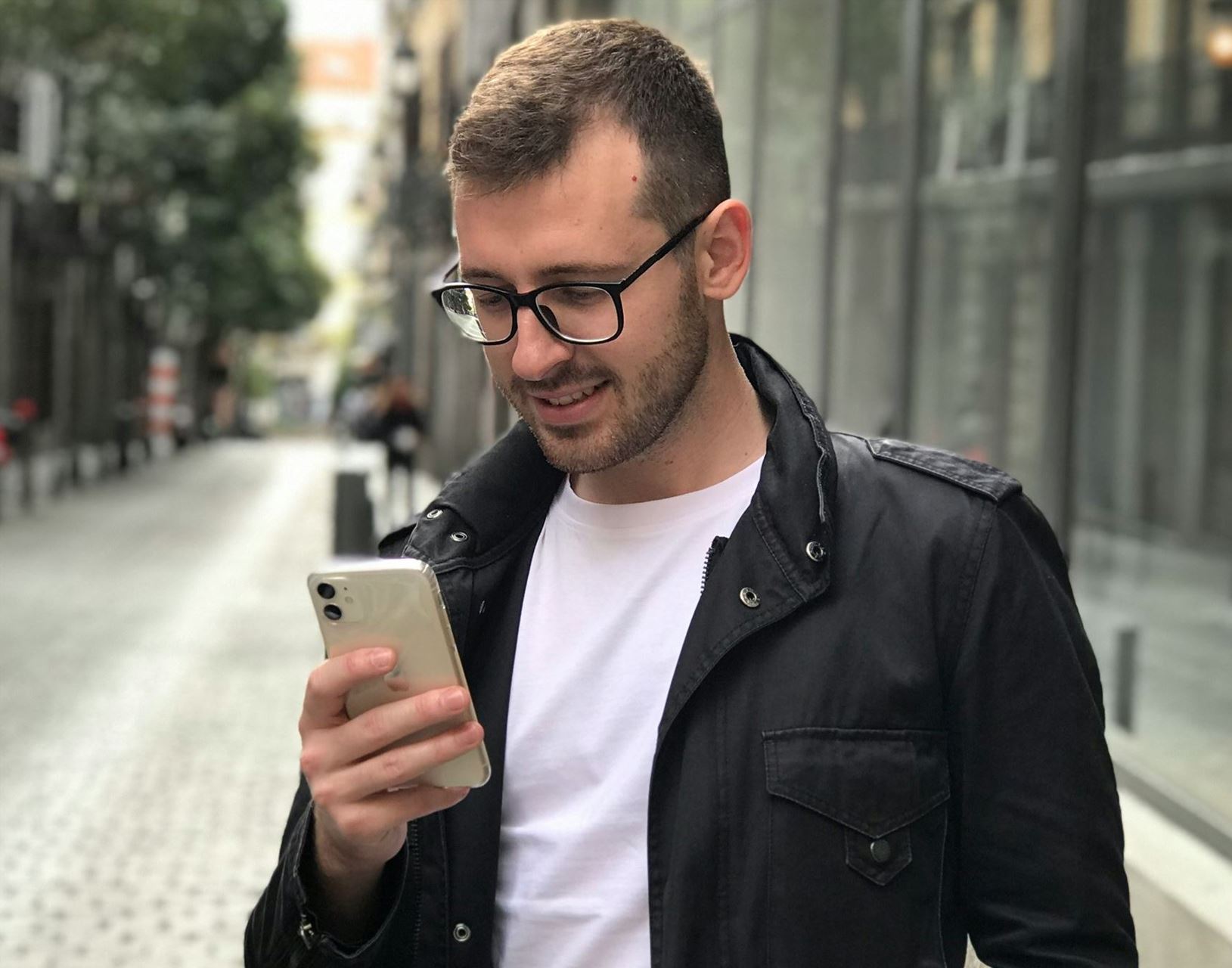 a young man with glasses wearing a black jacket is looking at his mobile phone.  He is stood in the street of a shopping precicnt